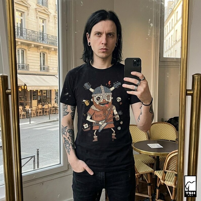 May include: Black t-shirt featuring a cartoon Viking playing a guitar, with the word "RAID" on the shirt. The image also shows a person wearing the shirt, black jeans, and holding a phone. The background includes a mirror and a window.