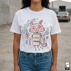 Pu&ograve; includere: T-shirt bianca con un disegno di gatto dei cartoni animati. Il gatto ha la testa e il corpo rosa, che tiene in mano un cartone di latte con schizzi d'acqua. Il corpo del gatto &egrave; a forma di scatola con la scritta "LUCK".
