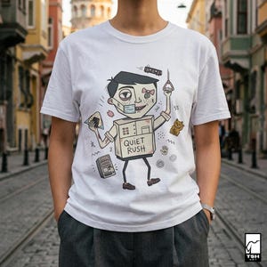 Op de afbeelding: Wit t-shirt met een cartoon illustratie. Het ontwerp toont een personage met een doosvormig lichaam, die een treinhandvat en een snack vasthoudt, met de tekst "QUIET RUSH" en andere grafische elementen.