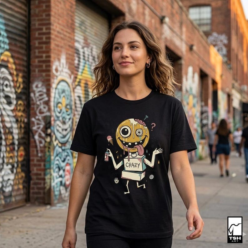 May include: Black t-shirt with a cartoon character design. The character has a yellow head, a brain, and a long tongue. The word "CRAZY" is printed on the character's body. The background shows graffiti art and a brick wall.
