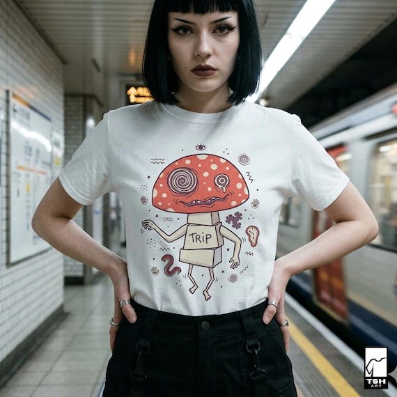 May include: White t-shirt featuring a cartoon mushroom character with a red cap and white spots. The mushroom has a face with swirling eyes and the word "TRIP" on its body. The shirt is worn by a person in a subway setting.