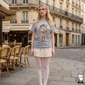 May include: A gray t-shirt with a cartoon illustration of a French character wearing a beret, striped shirt, and trench coat. The character is holding a cigarette and has the Eiffel Tower on their head. The shirt is paired with a cream-colored pleated skirt.