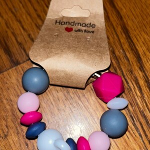 K&ouml;nnte beinhalten: Ein handgemachtes Bei&szlig;armband mit einer Vielzahl farbiger Perlen. Das Armband hat graue, rosa und pinkfarbene Perlen sowie ein wei&szlig;es Bei&szlig;element. Auf dem Anh&auml;nger steht "Handmade with love."