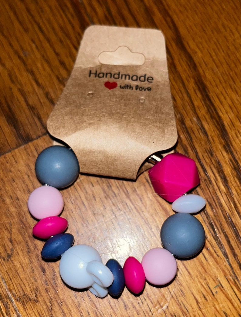 K&ouml;nnte beinhalten: Ein handgemachtes Bei&szlig;armband mit einer Vielzahl farbiger Perlen. Das Armband hat graue, rosa und pinkfarbene Perlen sowie ein wei&szlig;es Bei&szlig;element. Auf dem Anh&auml;nger steht "Handmade with love."