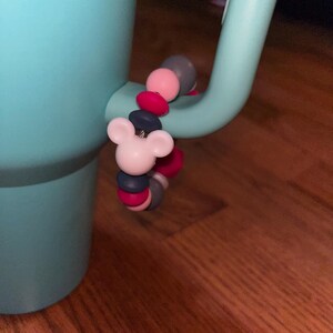 K&ouml;nnte beinhalten: Ein hellblauer Becher mit einem dekorativen Anh&auml;nger am Griff. Der Anh&auml;nger zeigt einen wei&szlig;en Mickey Mouse-Kopf und eine Reihe bunter Perlen in Rosa, Grau und Marineblau. Der Becher steht auf einer Holzoberfl&auml;che.