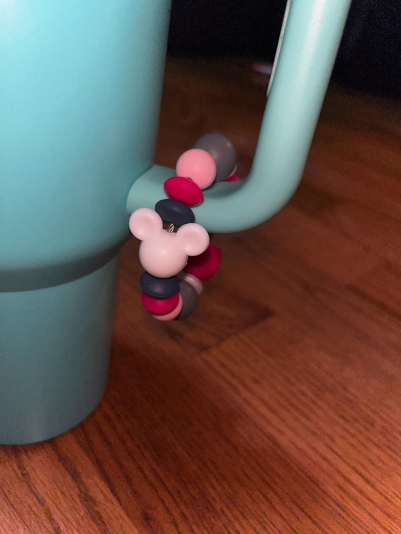 K&ouml;nnte beinhalten: Ein hellblauer Becher mit einem dekorativen Anh&auml;nger am Griff. Der Anh&auml;nger zeigt einen wei&szlig;en Mickey Mouse-Kopf und eine Reihe bunter Perlen in Rosa, Grau und Marineblau. Der Becher steht auf einer Holzoberfl&auml;che.