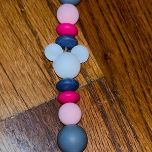 K&ouml;nnte beinhalten: Ein farbenfroher Schnullerclip mit einer Vielzahl von Perlen in Rosa, Blau, Grau und Wei&szlig;. Der Clip hat ein Mickey Mouse-Kopf-Design und ein geometrisches rosa Endst&uuml;ck.