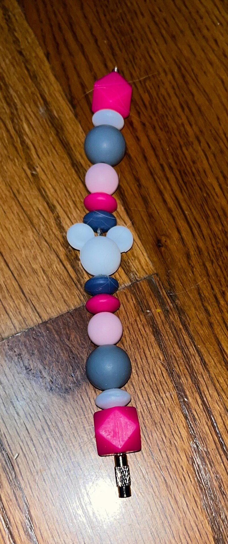 K&ouml;nnte beinhalten: Ein farbenfroher Schnullerclip mit einer Vielzahl von Perlen in Rosa, Blau, Grau und Wei&szlig;. Der Clip hat ein Mickey Mouse-Kopf-Design und ein geometrisches rosa Endst&uuml;ck.