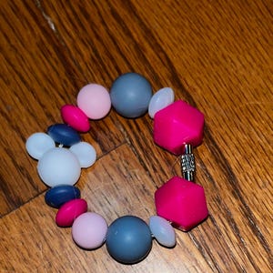 K&ouml;nnte beinhalten: Ein farbenfrohes Armband mit Perlen in Rosa, Blau, Grau und Wei&szlig;. Das Armband hat runde, ovale und geometrische Perlen, die durch einen silberfarbenen Verschluss verbunden sind. Die Perlen scheinen aus einem glatten, m&ouml;glicherweise Silikonmaterial zu bestehen.