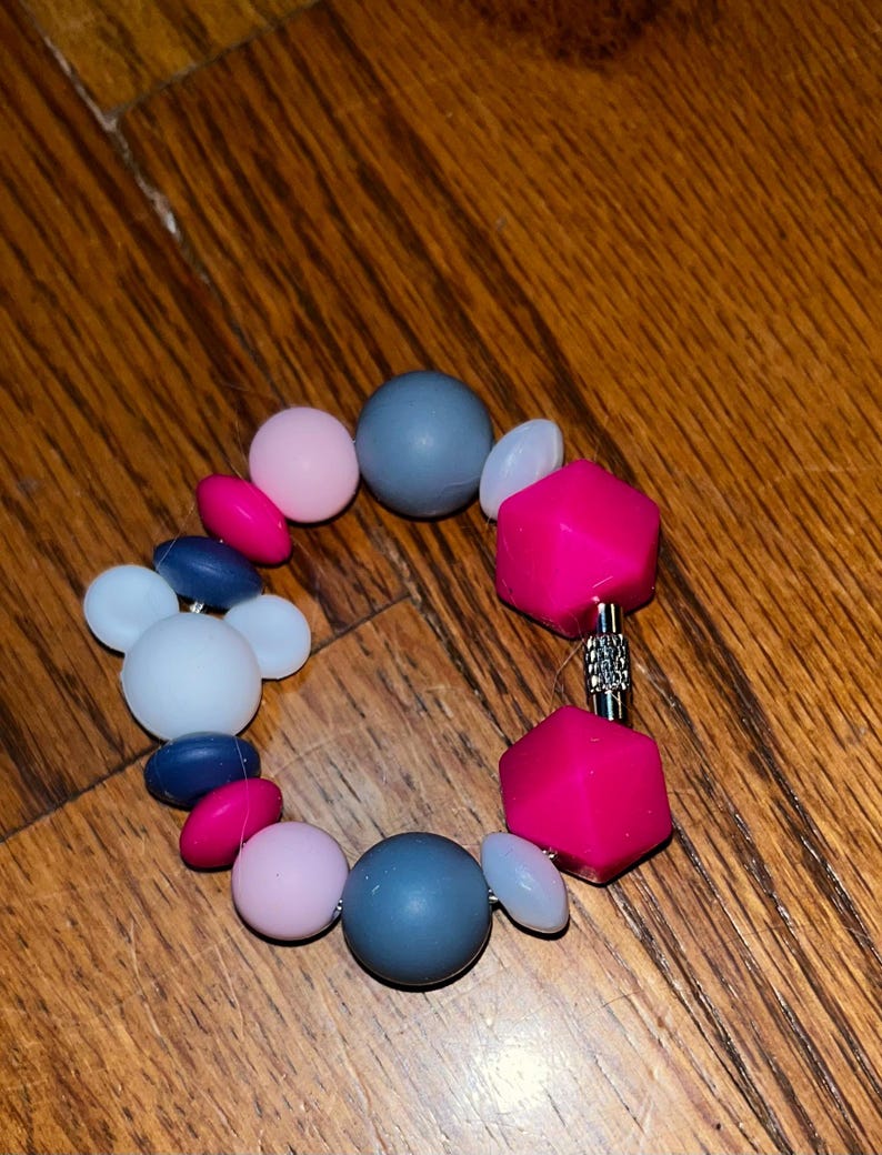 K&ouml;nnte beinhalten: Ein farbenfrohes Armband mit Perlen in Rosa, Blau, Grau und Wei&szlig;. Das Armband hat runde, ovale und geometrische Perlen, die durch einen silberfarbenen Verschluss verbunden sind. Die Perlen scheinen aus einem glatten, m&ouml;glicherweise Silikonmaterial zu bestehen.