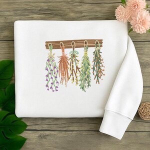 May include: White sweatshirt featuring an embroidered design of hanging herbs. The design includes various herbs in shades of green, purple, and orange, suspended from a wooden beam. The sweatshirt has a crew neck and long sleeves.