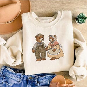 May include: Cream-colored sweatshirt featuring an embroidered design of two bears holding hands. The bear on the left wears overalls and a cap, while the bear on the right wears a bonnet and apron. The sweatshirt is paired with blue denim shorts and a brown hat.