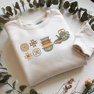 May include: Cream-colored sweatshirt with a crew neck and long sleeves. The front features a colorful graphic of stacked bowls, a teapot, teacup, and geometric shapes in shades of green, orange, and brown. The sweatshirt is folded on a white surface.