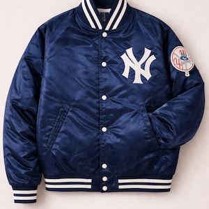 Pode incluir: Casaco bomber de cetim azul marinho com detalhes brancos e fecho de botão branco. O casaco apresenta o logotipo dos New York Yankees no peito e um patch da equipa na manga. A gola e os punhos têm riscas brancas.