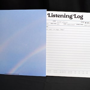 May include: A listening log notebook with a light blue cover featuring a rainbow design. The notebook is open, revealing lined pages and a header that reads "Listening Log". The notebook is on a black surface.