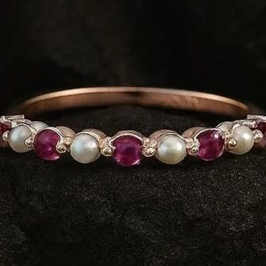 May include: A rose gold band ring featuring alternating round, vibrant red gemstones and white pearls. The ring is set against a dark, textured background, highlighting the contrast of colors and the ring's delicate design. A piece of jewelry.