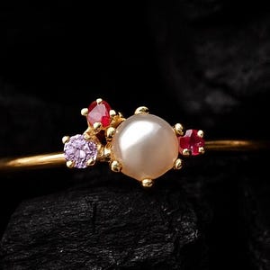Może przedstawiać: Złoty pierścionek z dużą, okrągłą perłą otoczoną małymi czerwonymi i fioletowymi kamieniami szlachetnymi. Pierścionek jest osadzony na ciemnym, teksturowanym tle, podkreślając delikatny design i kontrast kolorystyczny biżuterii.