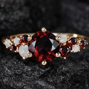 Może przedstawiać: Złoty pierścionek z dużym, okrągłym, ciemnoczerwonym kamieniem szlachetnym w centrum, otoczonym mniejszymi czerwonymi i białymi kamieniami. Pierścionek jest umieszczony na ciemnym, teksturowanym tle, podkreślającym blask kamieni.