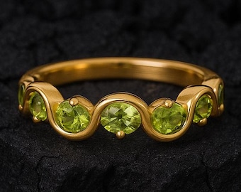 Könnte beinhalten: Ein goldener Ring mit einer Reihe von oval geschliffenen, hellgrünen Edelsteinen. Das Band des Rings hat ein wellenförmiges Design, wobei jeder Edelstein in einem separaten, gebogenen Abschnitt gefasst ist. Der Ring wird vor einem dunklen, strukturierten Hintergrund präsentiert.