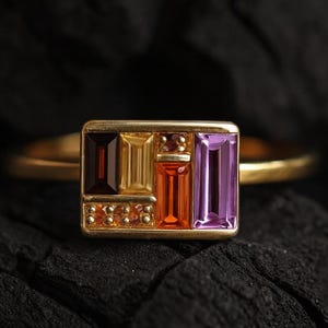 Op de afbeelding: Een gouden ring met een rechthoekige voorkant met kleurrijke edelstenen. De edelstenen zijn in tinten rood, oranje, geel en paars. De ring wordt getoond tegen een donkere, getextureerde achtergrond.
