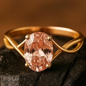 Può includere: Un anello d'oro con una gemma ovale color pesca. La fascia dell'anello presenta un design intrecciato e la gemma è tenuta in posizione da quattro piccole griffe d'oro. L'anello è posto su uno sfondo scuro e strutturato.