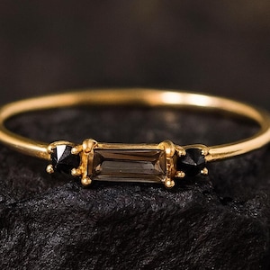 Peut inclure: Une bague en or avec une pierre précieuse rectangulaire marron au centre, flanquée de deux petites pierres précieuses noires. La bague est posée sur une surface sombre et texturée, mettant en valeur son design délicat et son élégance.