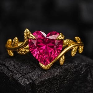 May include: A gold ring featuring a heart-shaped, vibrant pink gemstone. The ring's band is adorned with delicate leaf-like designs, creating an elegant and romantic aesthetic. The gemstone is set in a gold bezel.
