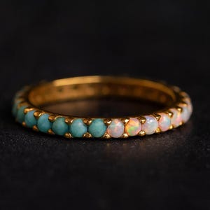 Può includere: Un anello d'oro con una fascia di pietre preziose turchesi e opali alternate. Le pietre turchesi sono di colore teal, mentre gli opali presentano tonalità pastello. L'anello è posto su uno sfondo scuro.