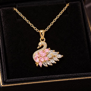 May include: A gold-toned necklace featuring a swan-shaped pendant adorned with marquise-cut pink and clear crystals. The pendant hangs from a delicate gold chain inside a black jewellery box with the text Twinkletrailstudio printed in gold.