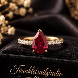 Può includere: Un anello in oro con una grande pietra rossa taglio a goccia come pietra centrale, affiancata da una fila di piccole pietre chiare taglio rotondo lungo la fascia. L'anello poggia su un cofanetto in velluto nero con il testo dorato Twinkletrailstudio.