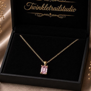 May include: A gold necklace with a rectangular pink gemstone pendant, presented in a black jewelry box. The box is open, revealing the necklace against a black interior. The text "Twinkletrailstudio" is displayed on the inside of the box.