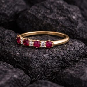 May include: A gold band ring set with alternating red gemstones and clear, round-cut stones. The ring is displayed on a dark, textured surface, highlighting the sparkle of the gemstones. The ring is a piece of jewelry.
