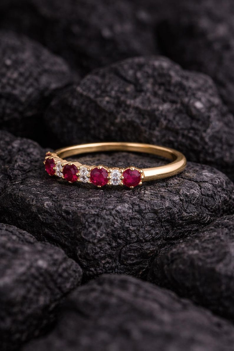 May include: A gold band ring set with alternating red gemstones and clear, round-cut stones. The ring is displayed on a dark, textured surface, highlighting the sparkle of the gemstones. The ring is a piece of jewelry.