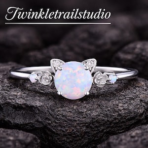 May include: A silver ring featuring a large, round, iridescent opal center stone. The ring has cat ear accents with small clear stones and floral details on the band. The text "Twinkletrailstudio" is at the top.