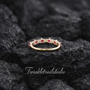 May include: A gold ring featuring a row of small, round, vibrant red rubies set along the top band. The ring is positioned horizontally, showcasing the metal band and the gemstone setting. The text Twinkletrailstudio is visible.