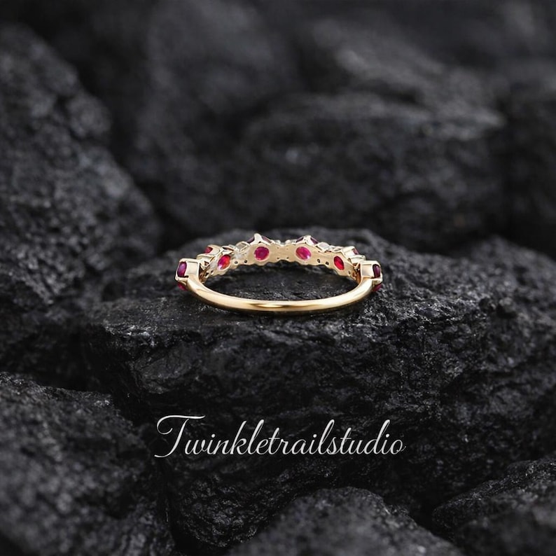 May include: A gold ring featuring a row of small, round, vibrant red rubies set along the top band. The ring is positioned horizontally, showcasing the metal band and the gemstone setting. The text Twinkletrailstudio is visible.