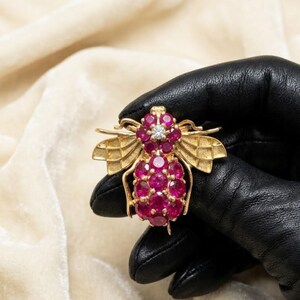 Puede incluir: Broche de abeja dorada con detalles de rubíes y diamantes. El cuerpo de la abeja está compuesto por numerosos rubíes redondos de color rojo vibrante. Las alas son detalladas y texturizadas. Un solo diamante está engastado en la parte superior de la cabeza de la abeja.