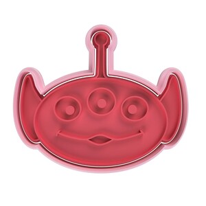 May include: A red and pink cookie cutter in the shape of a three-eyed alien with a smiling face. The cookie cutter has a rounded shape with a small antenna on top and large ears on the sides.