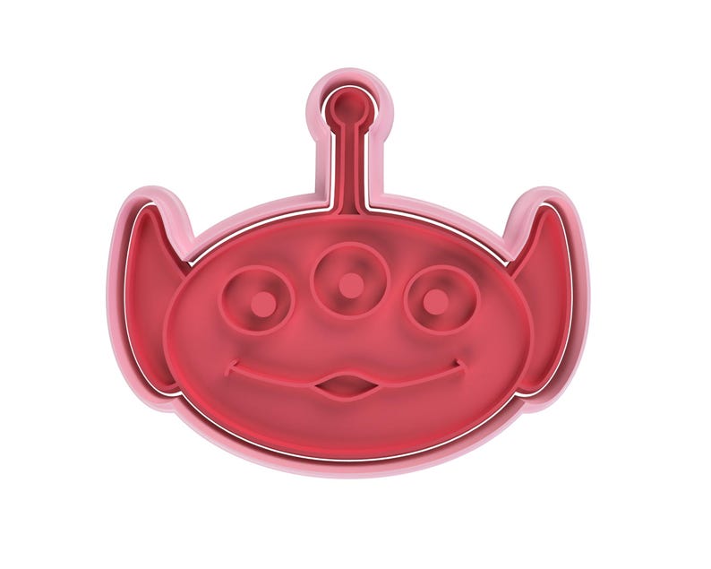 May include: A red and pink cookie cutter in the shape of a three-eyed alien with a smiling face. The cookie cutter has a rounded shape with a small antenna on top and large ears on the sides.