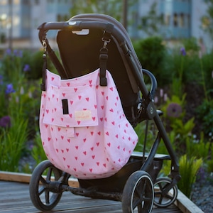Puede incluir: Una bolsa organizadora rosa para cochecito de bebé con un patrón de corazones rojos. La bolsa está sujeta a un cochecito negro. El fondo incluye vegetación y un edificio.