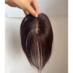 May include: A dark brown hair topper, held up against a white background. The hairpiece has a natural-looking part and appears to be made of real hair. The hair is a rich, warm brown color.