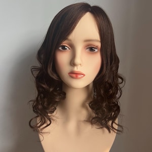 May include: A mannequin head with a brown wig featuring a side part and shoulder-length, wavy curls. The wig has a natural-looking hairline and is styled with a slight wave. The mannequin has a fair complexion, with painted eyes and lips.