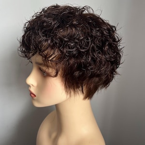 May include: A short, dark brown wig with tight, defined curls and a layered cut. The hair texture appears wet or styled with product, creating a glossy finish. The wig is displayed on a smooth, neutral-toned mannequin head, showing the profile view.