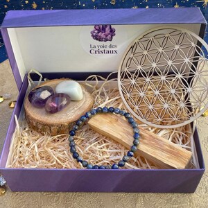 Peut inclure: Une boîte cadeau violette contenant un bracelet de perles bleues, un disque en bois avec un motif fleur de vie découpé au laser, une tranche de bois avec deux améthystes polies et une pierre blanche, et un bâtonnet, avec l'étiquette La voie des Cristaux.