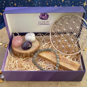 Peut inclure: Une boîte cadeau violette contenant une tranche de bois ronde avec une pierre rose, blanche et violette, un bracelet de perles grises, un bâton de bois et une découpe en bois en forme de fleur de vie. L'autocollant indique La voie des Cristaux.