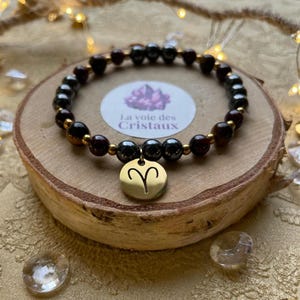 Könnte beinhalten: Ein Perlenarmband mit abwechselnd dunkelbraunen und metallisch grauen, kugelförmigen Perlen und kleinen goldfarbenen Zwischenelementen. Ein runder, goldfarbener Anhänger mit dem eingravierten Widder-Sternzeichen hängt in der Mitte.