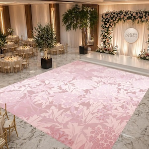 May include: A large, light pink floral patterned dance floor dominates the center of a ballroom. The floor is surrounded by round tables with gold chairs, and a stage decorated with flowers. The words "Gloria Designs" are on the stage.