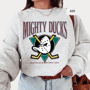 May include: Light grey sweatshirt featuring the text "MIGHTY DUCKS" in maroon lettering above a teal and black graphic of a hockey mask and crossed hockey sticks. The text "INAUGURAL SEASON 1993-1994" is below the graphic.