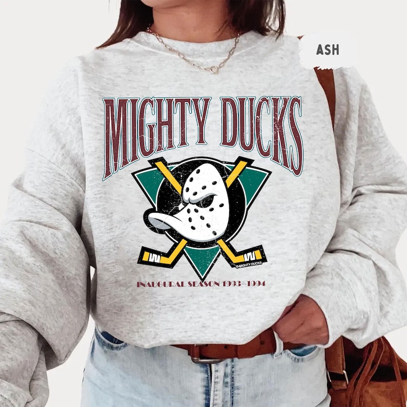 May include: Light grey sweatshirt featuring the text "MIGHTY DUCKS" in maroon lettering above a teal and black graphic of a hockey mask and crossed hockey sticks. The text "INAUGURAL SEASON 1993-1994" is below the graphic.
