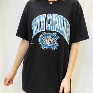 May include: Black t-shirt featuring the text "NORTH CAROLINA" in white and light blue arched lettering. Below the text is a light blue and white logo with a ram's head and the words "CAROLINA TAR HEELS".